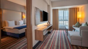 Habitación de hotel con cama, televisión, escritorio, silla y vistas a la ciudad a través de amplios ventanales.