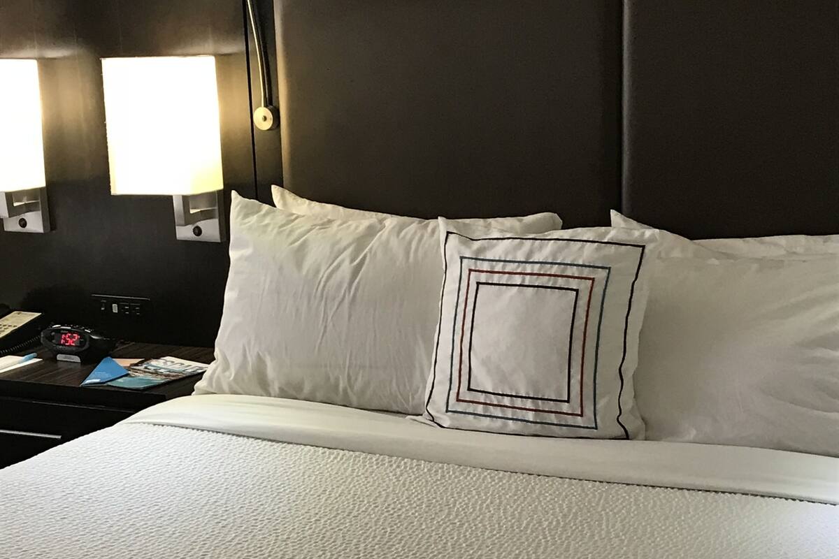 Un lit bien fait, avec une literie blanche et un coussin décoratif. On aperçoit une table de chevet avec une lampe et une horloge.
