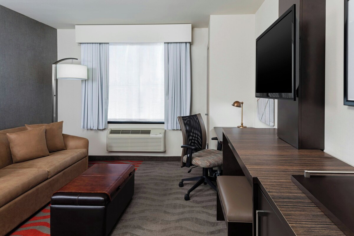 Ein Hotelzimmer mit einer Couch, einem Schreibtisch, einem Stuhl, einem Flachbildfernseher, einer Lampe und einem fenster mit Vorhängen.