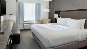 A hotel room with a large bed, a desk with a flat-screen TV, a chair, a lamp, and a window with curtains.