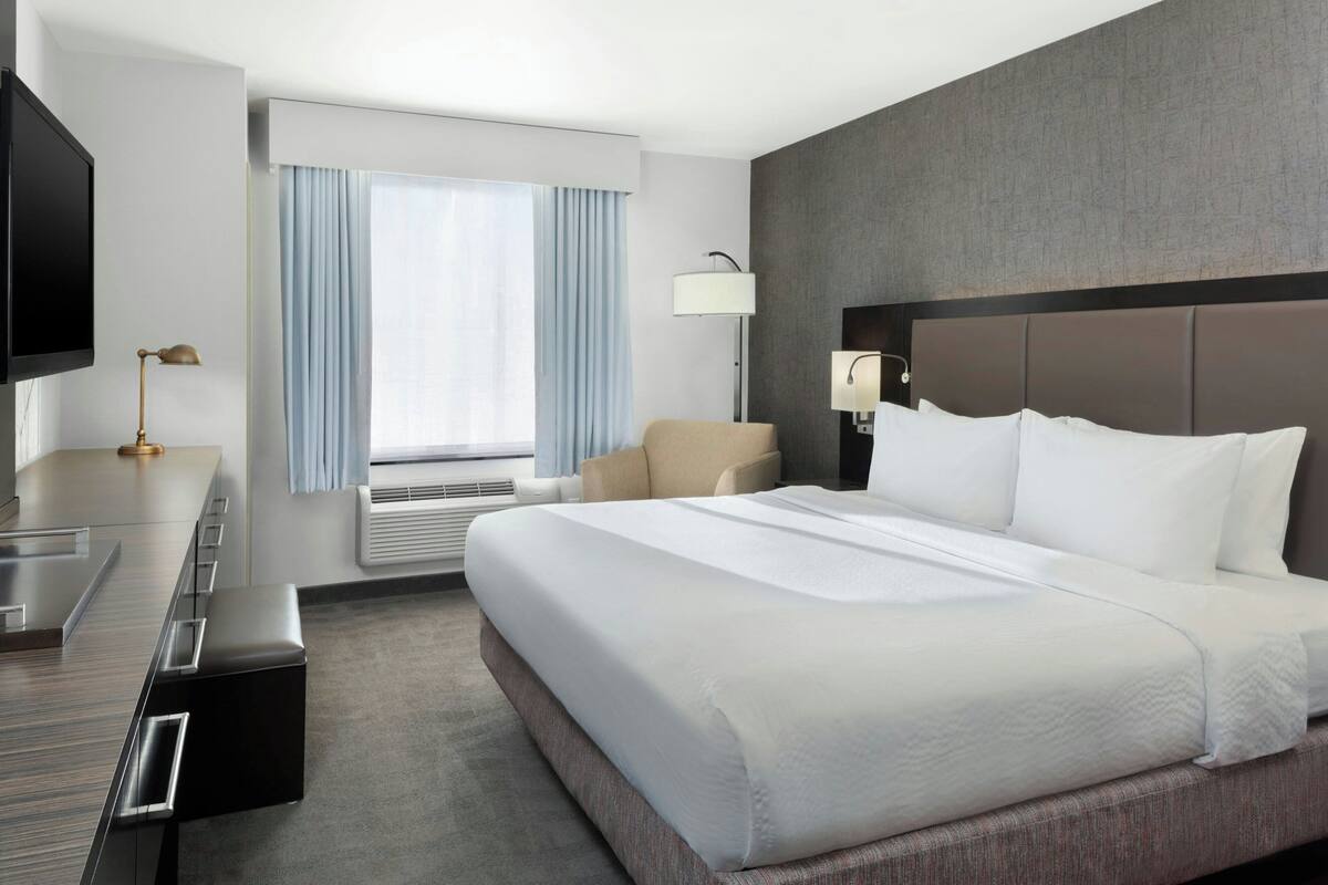 Une chambre d’hôtel avec un grand lit, un bureau doté d’un téléviseur à écran plat, une chaise, une lampe et une fenêtre avec des rideaux.