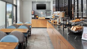 A modern cafe with wooden tables and chairs, a self-service counter with food items, and a television mounted on the wall.