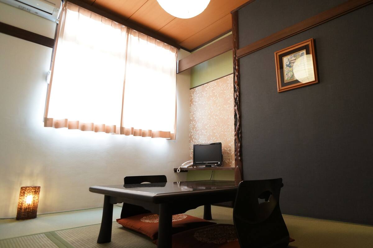 Ett traditionellt japanskt rum med tatamimattor på golvet, ett lågt bord, en stol, ett skrivbord med en dator och en inramad tavla på väggen.