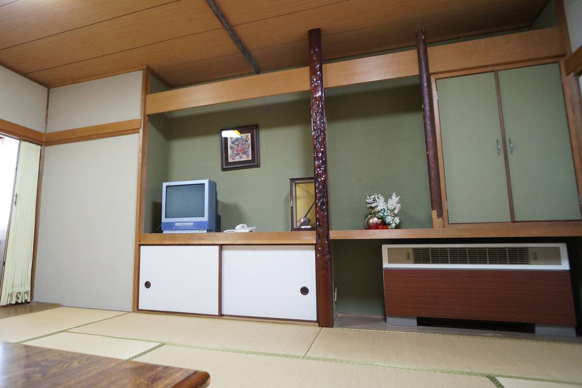 Ett traditionellt japanskt rum med tatamimattor på golvet, ett lågt träbord, en tv och ett skåp med en dekorativ figur.