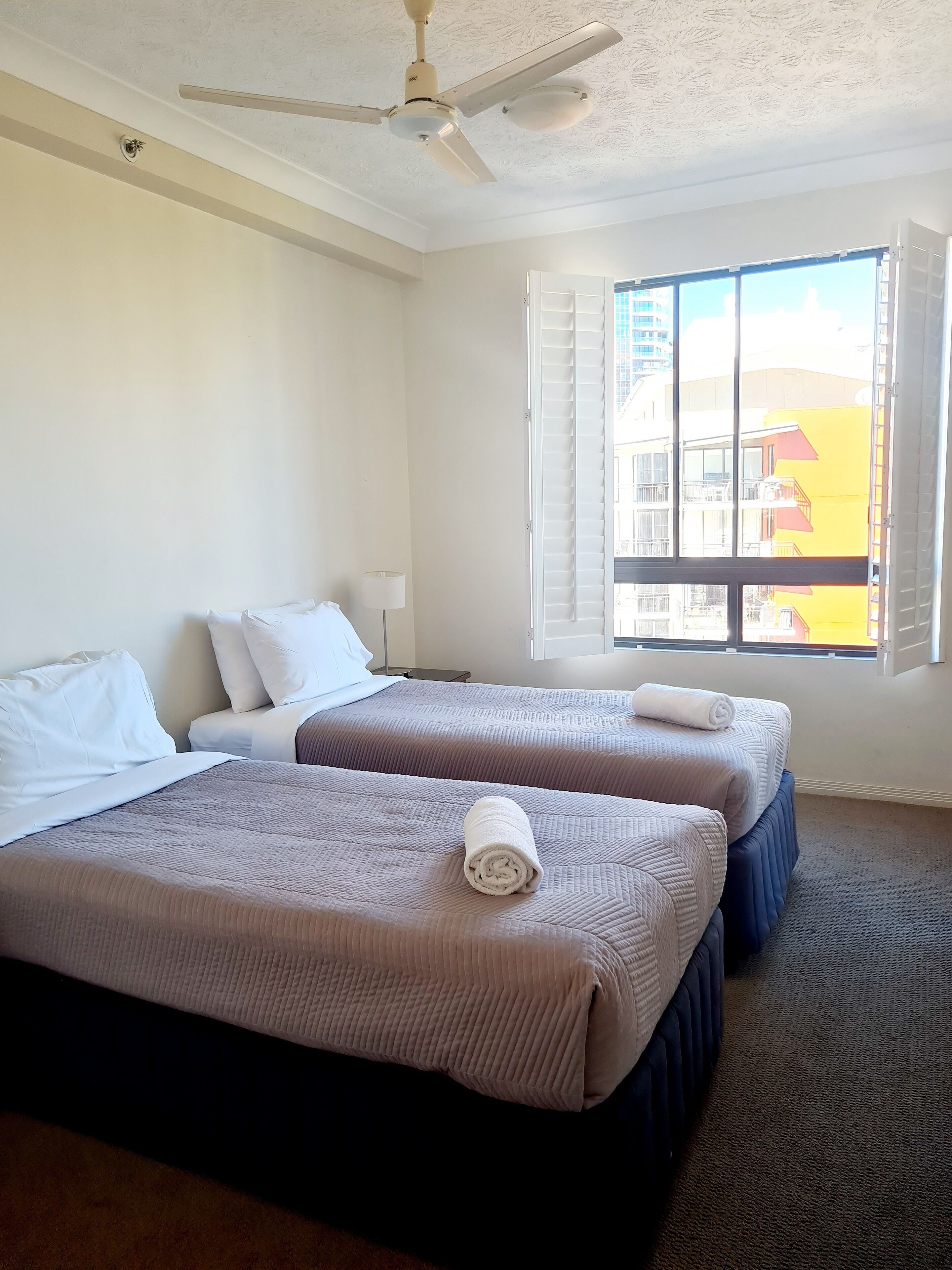 A hotel room with two beds, a ceiling fan, a window with blinds, and a view of a building outside.