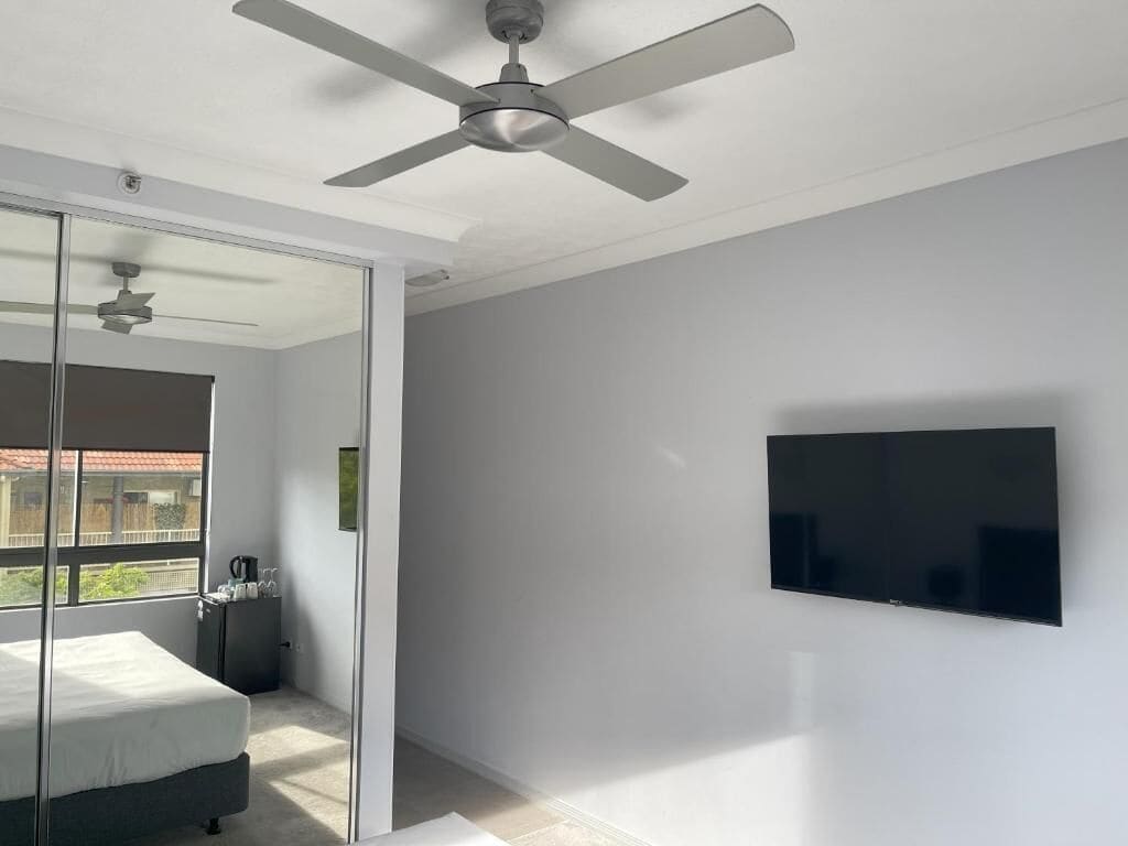 Une chambre moderne équipée d’un lit, d’un ventilateur de plafond, d’une télévision et d’une porte-fenêtre coulissante donnant sur un balcon.