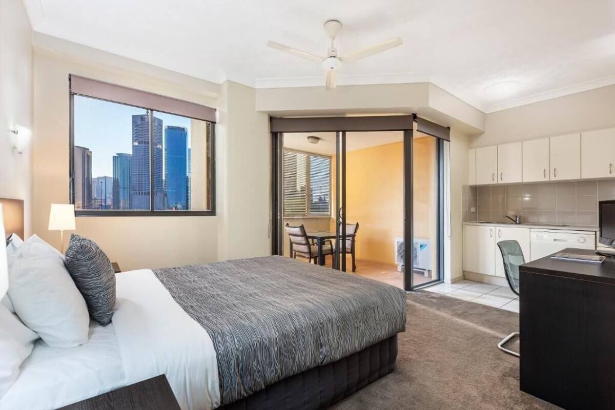 A hotel room with a bed, a desk, a chair, a city view, and a sliding door to a balcony.
