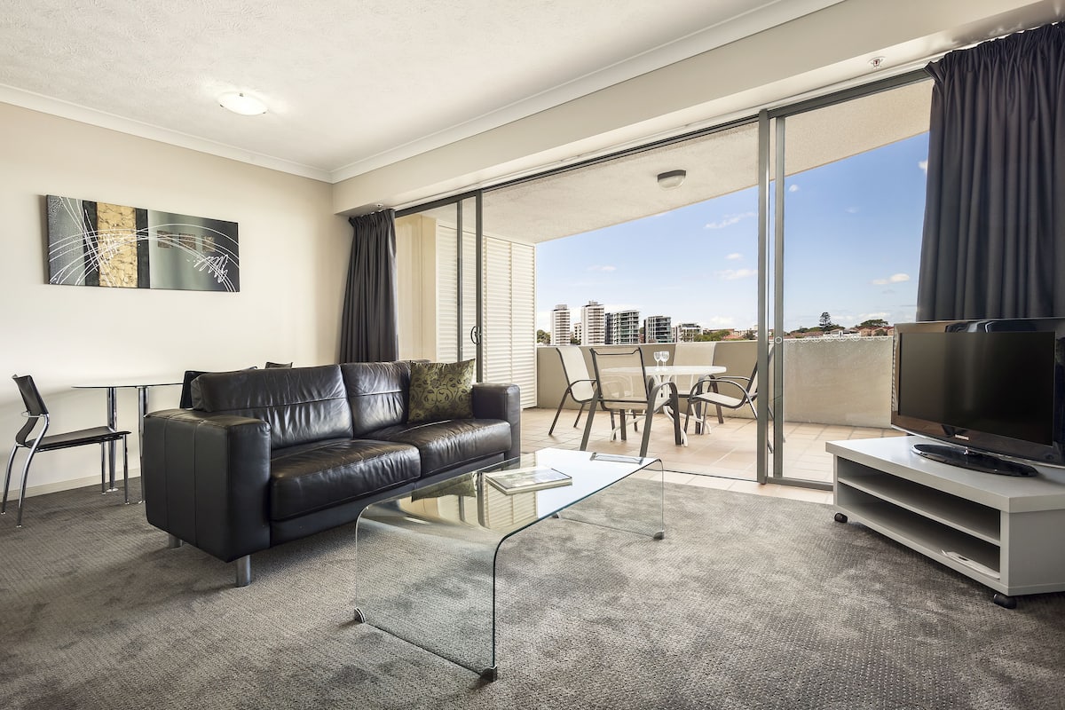 A modern living room with a black leather sofa, a glass coffee table, and a flat-screen TV on a white stand. There is a dining area with a table and chairs, and a balcony with a view of the city skyline.