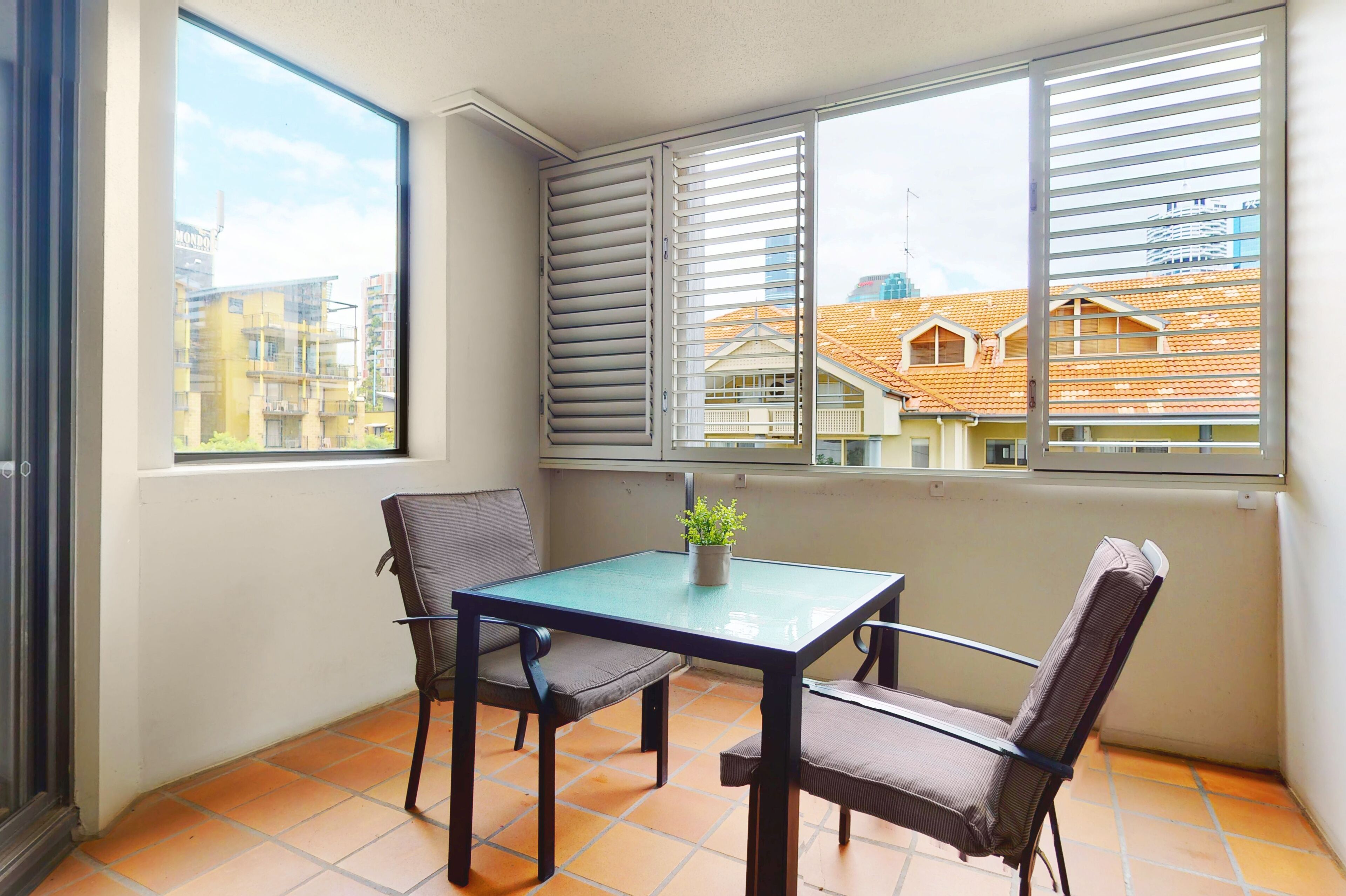 A balcony with a table and chairs, a potted plant, and a view of buildings through the window.