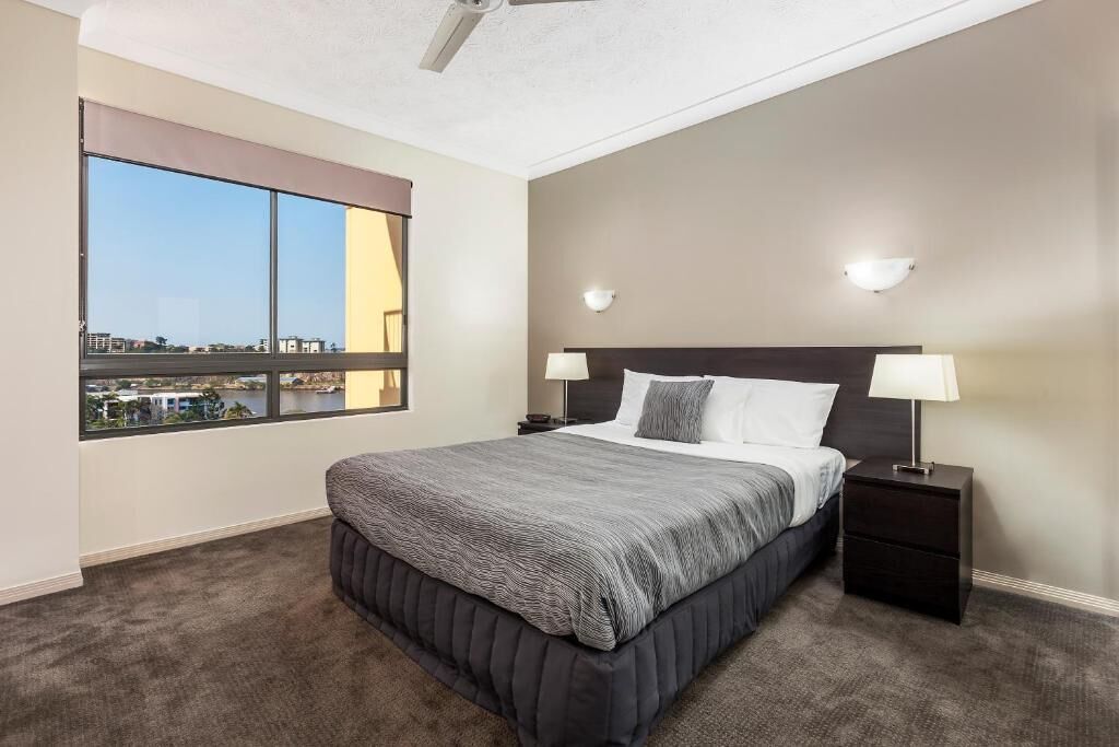 A bedroom with a large window, a bed with a grey bedspread, two bedside lamps, and a ceiling fan.