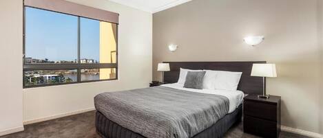A bedroom with a large window, a bed with a grey bedspread, two bedside lamps, and a ceiling fan.