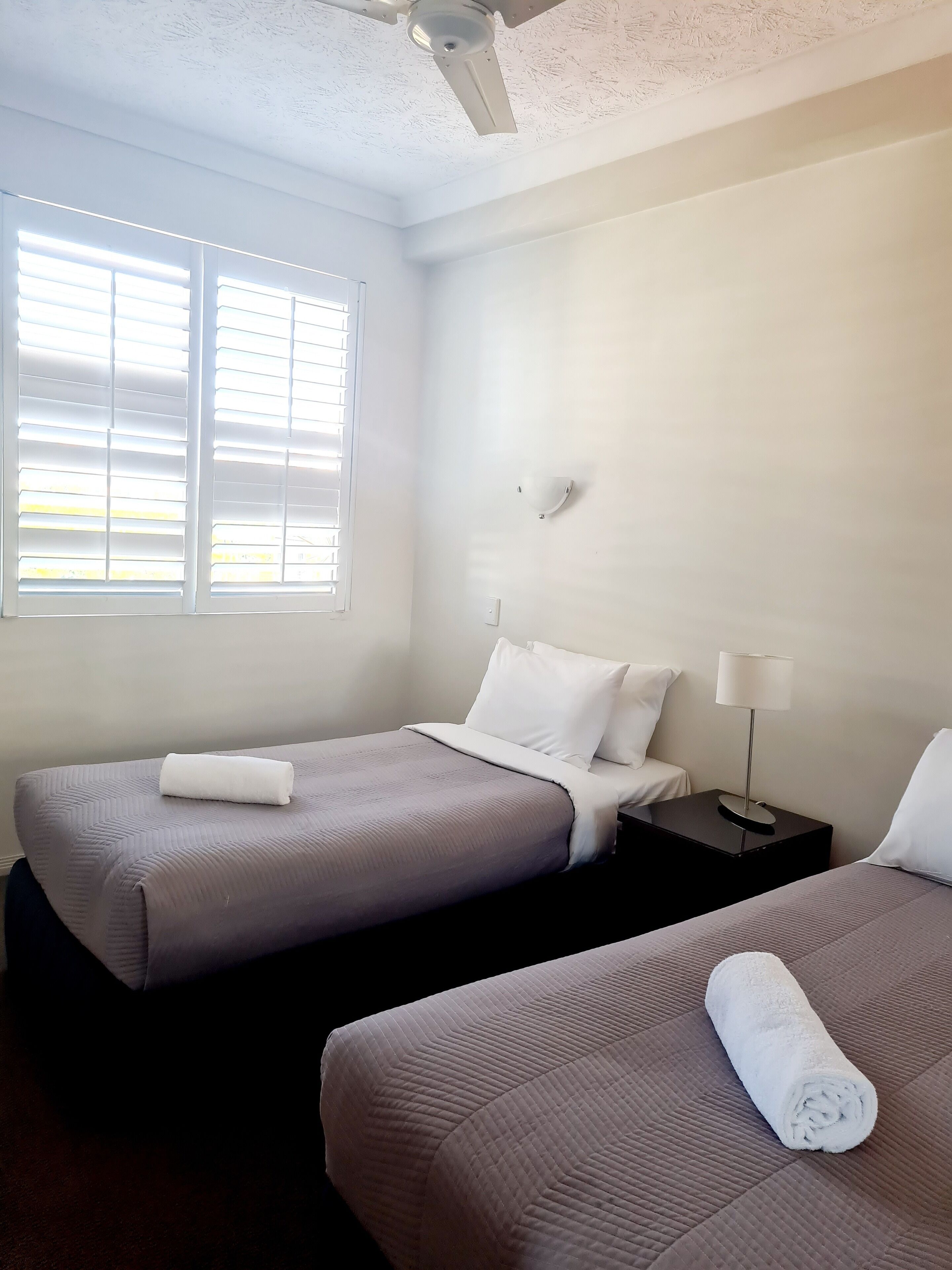 A hotel room with two single beds, a ceiling fan, a window with blinds, a wall-mounted light, and a nightstand with a lamp.