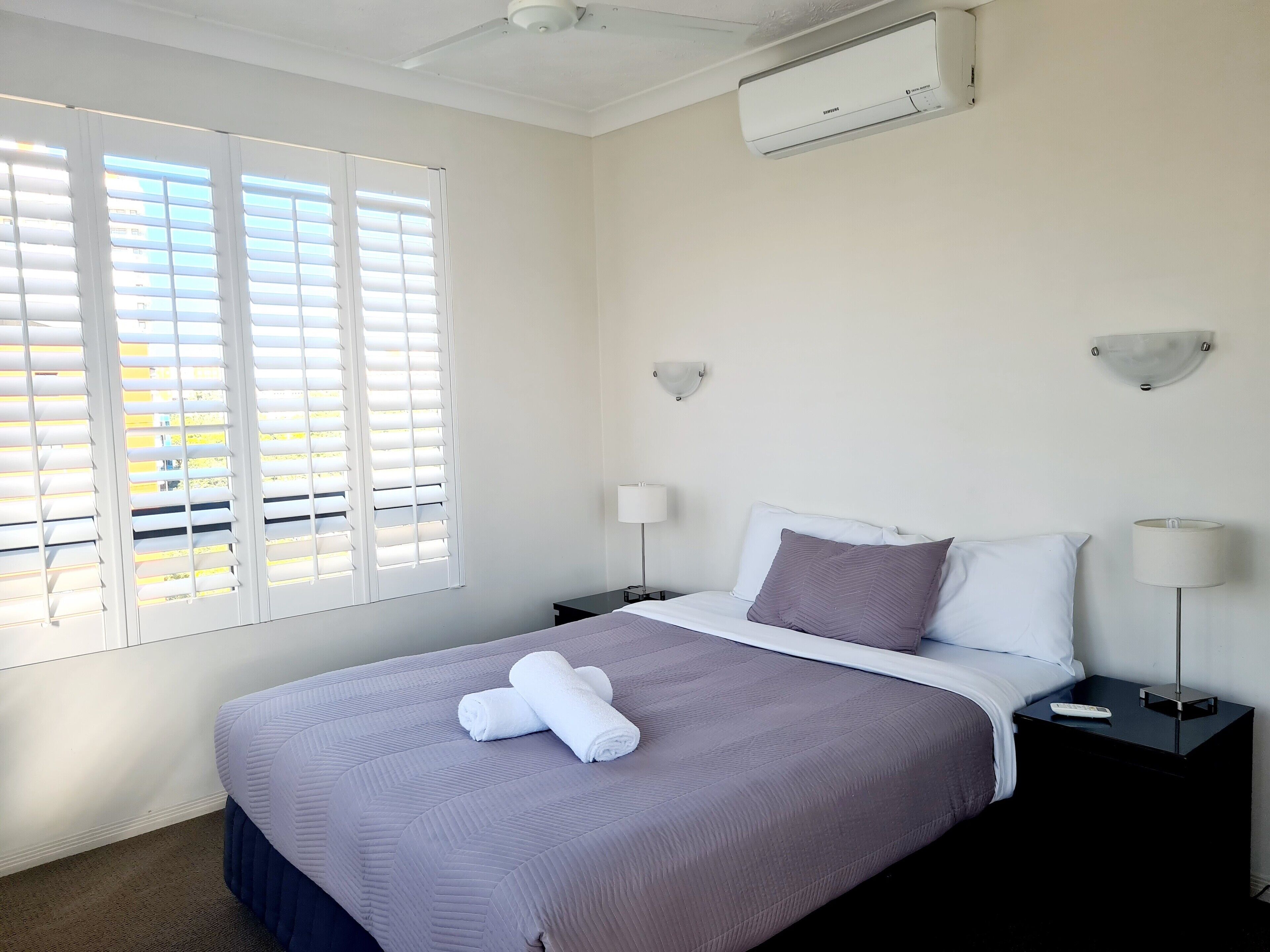 Habitación de hotel con cama, dos mesitas de noche, aire acondicionado instalado en la pared y una ventana con persianas.