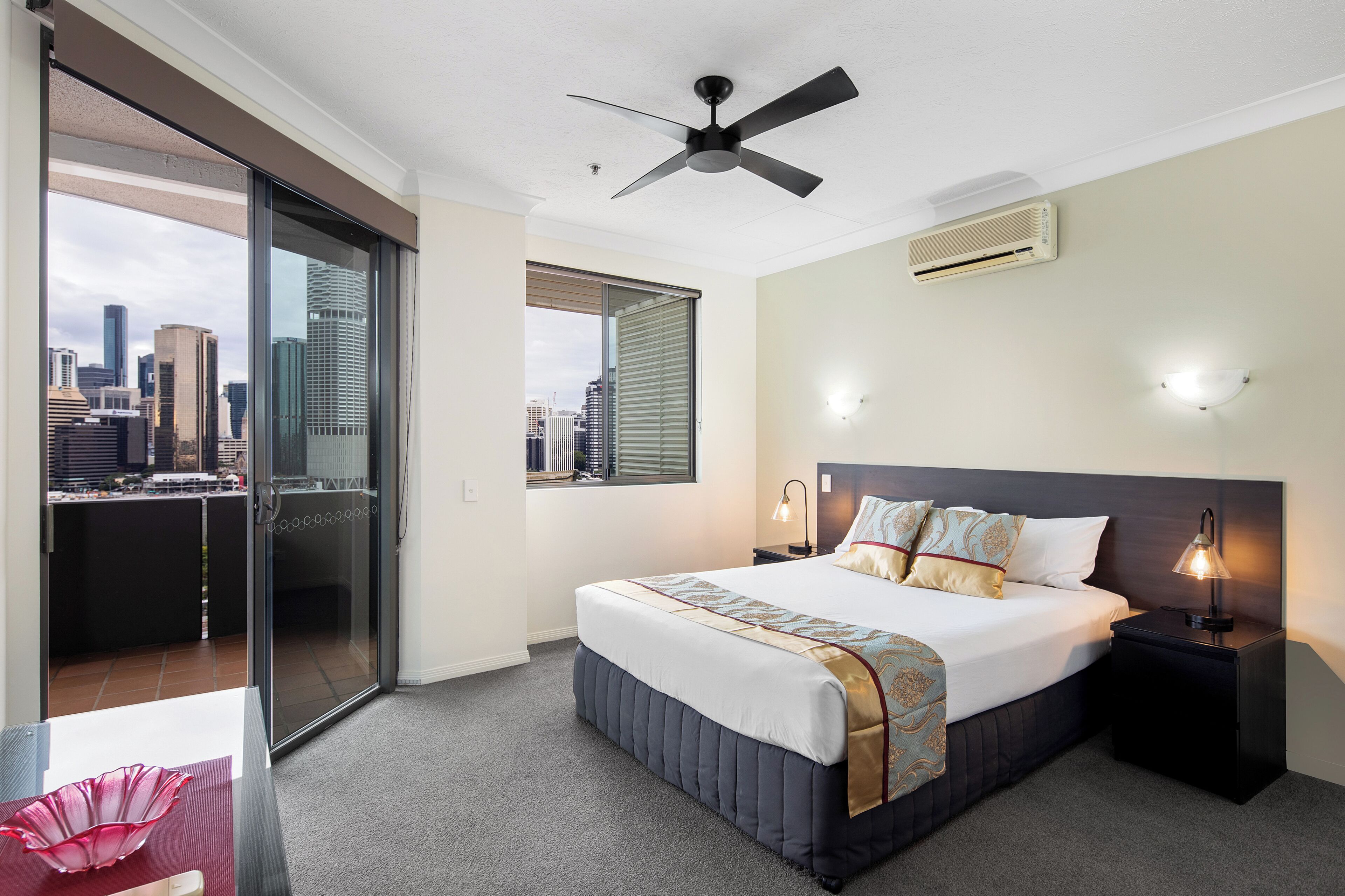 A hotel room with a bed, a nightstand, a ceiling fan, a window with a city view, and a sliding glass door.