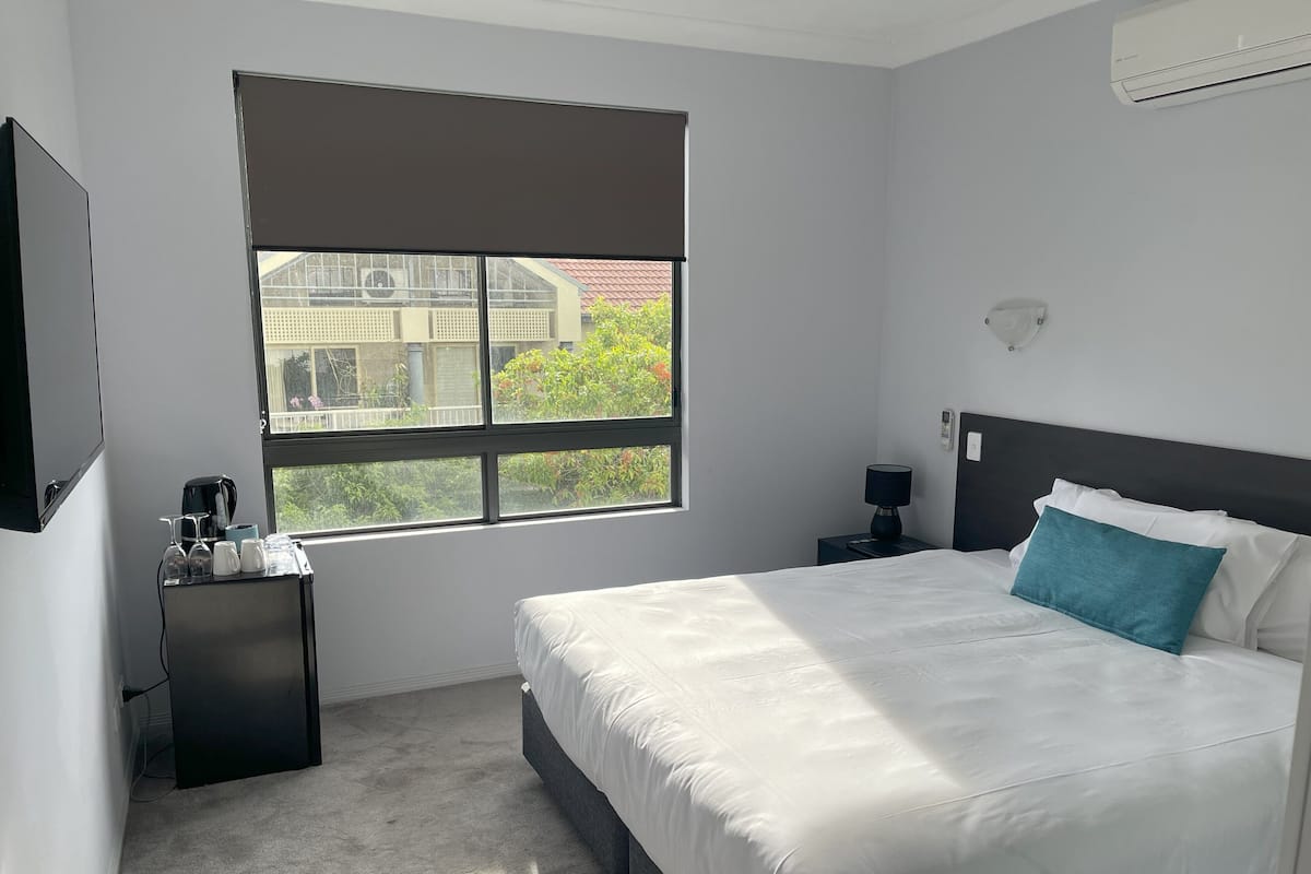A hotel room with a large bed, a nightstand, a desk with a coffee maker, and a window with a view of buildings and greenery.