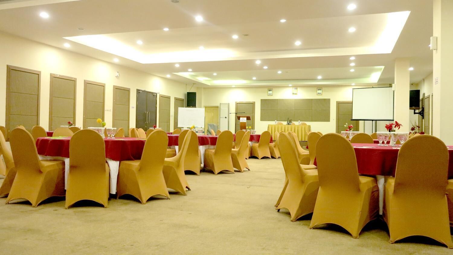 A conference room with rows of tables and chairs, equipped with a projector screen and a whiteboard.