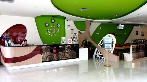A reception area with a modern design, including a green wall, a reception desk, and a digital screen. - Jolin Hotel (Makassar)
