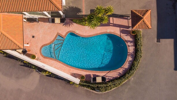 Vue aérienne d’une piscine à bordures courbes, entourée d’une terrasse en briques et de palmiers.