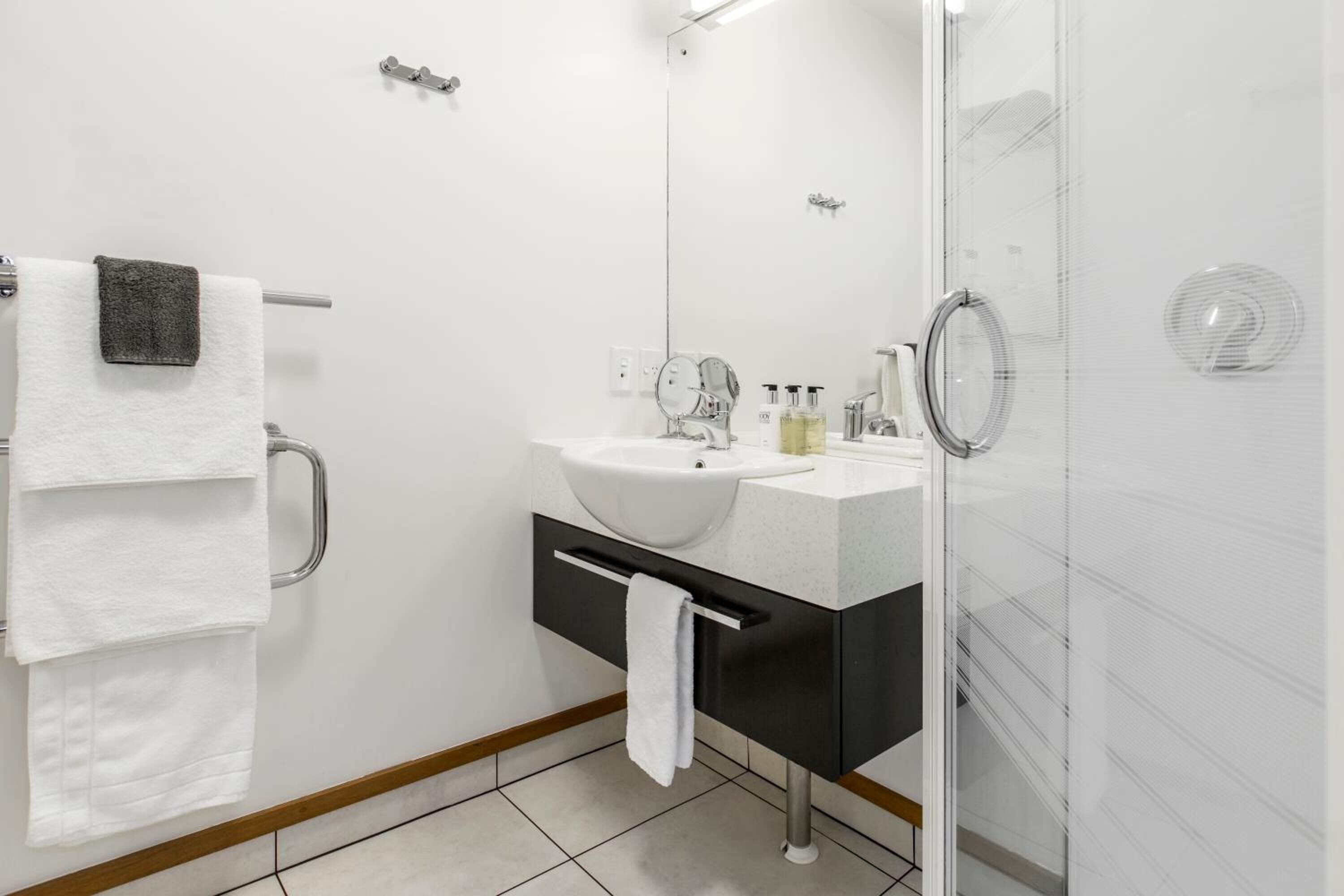 Un baño moderno con lavamanos blanco, tocador negro y cabina de ducha con mampara de vidrio.