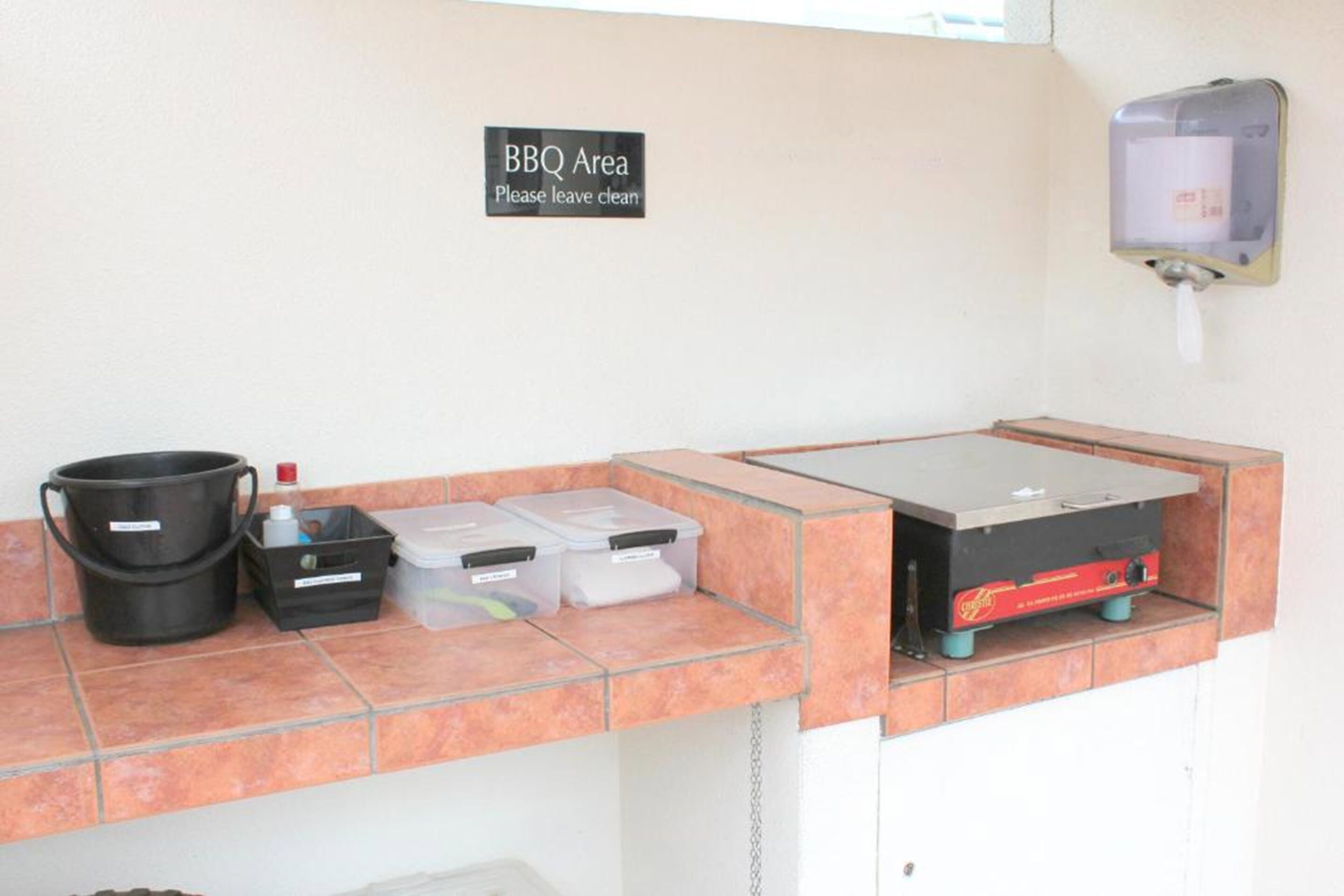 Een schone en geordende barbecueplek met een bord waarop staat te lezen om het schoon achter te laten. Er staan opslagbakken en een draagbare barbecuegrill op een betegelde toonbank.