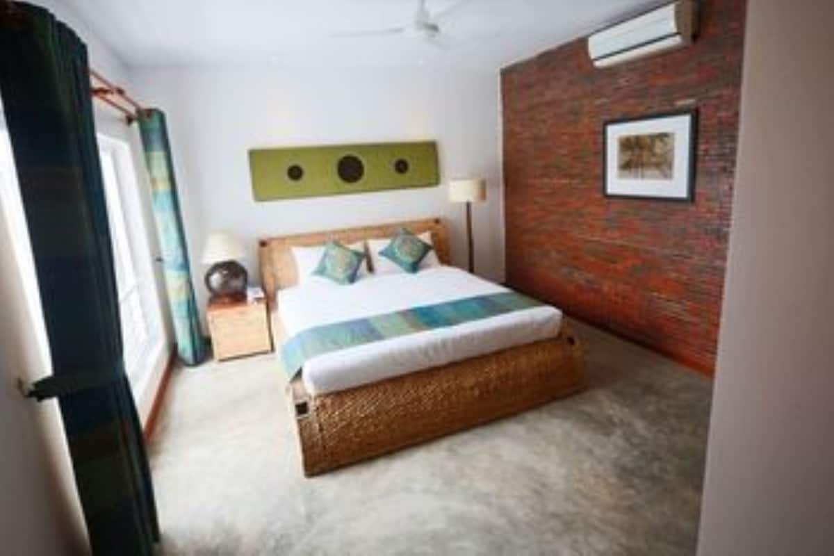 A bedroom with a bed, a headboard, a wall-mounted air conditioner, a lamp, and a picture frame.
