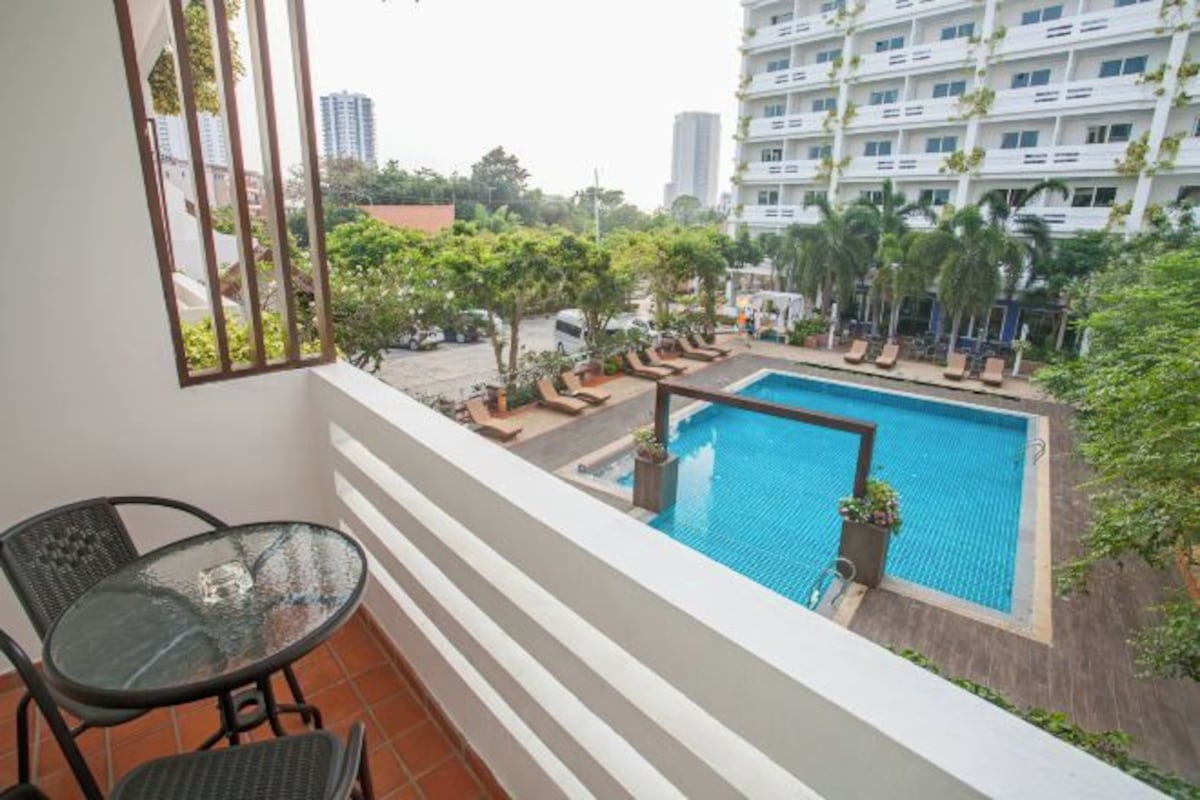 A balcony with a glass table and chairs, overlooking a swimming pool with lounge chairs and palm trees.