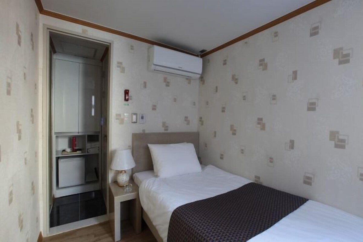 A hotel room with a bed, a desk, a chair, a wardrobe, and an air conditioning unit.