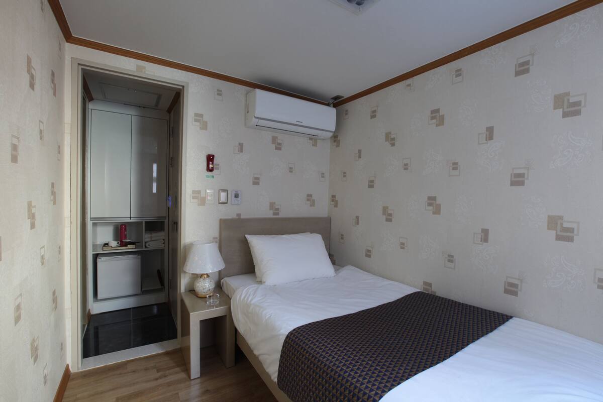 A hotel room with a bed, a nightstand with a lamp, a wardrobe, and an air conditioning unit on the wall.