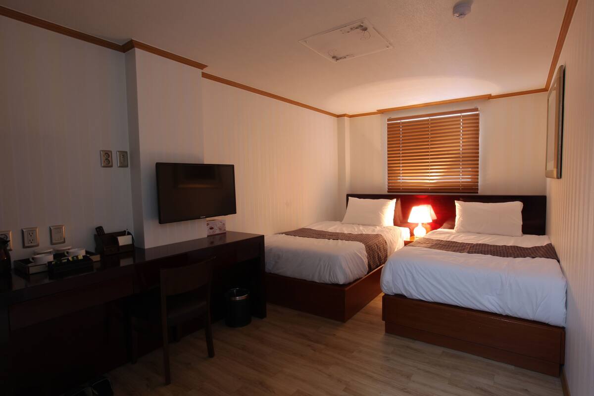 A hotel room with two beds, a desk, a television, and a window with blinds.