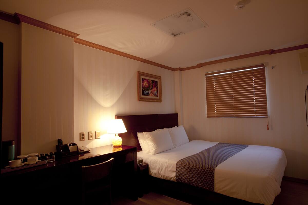 A hotel room with a bed, bedside table, lamp, and a window with blinds.
