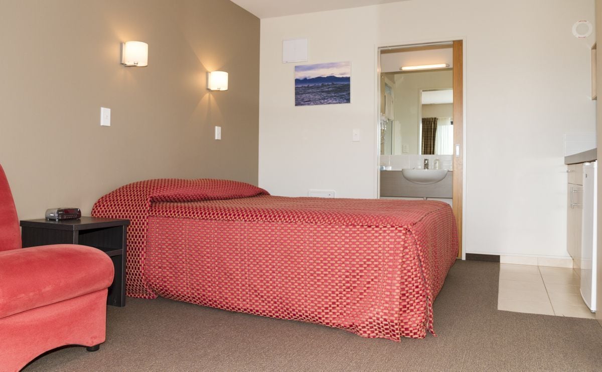 Une chambre d’hôtel avec un lit, une chaise rouge, un bureau avec une lampe, un miroir et une salle de bain visible à travers une porte ouverte.