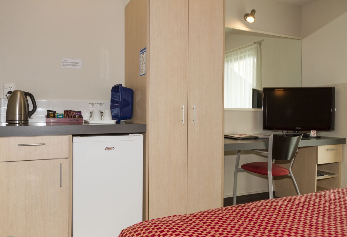 Habitación de hotel pequeña con kitchenette, una cama con cubrecama estampado, un escritorio con computadora y un televisor.