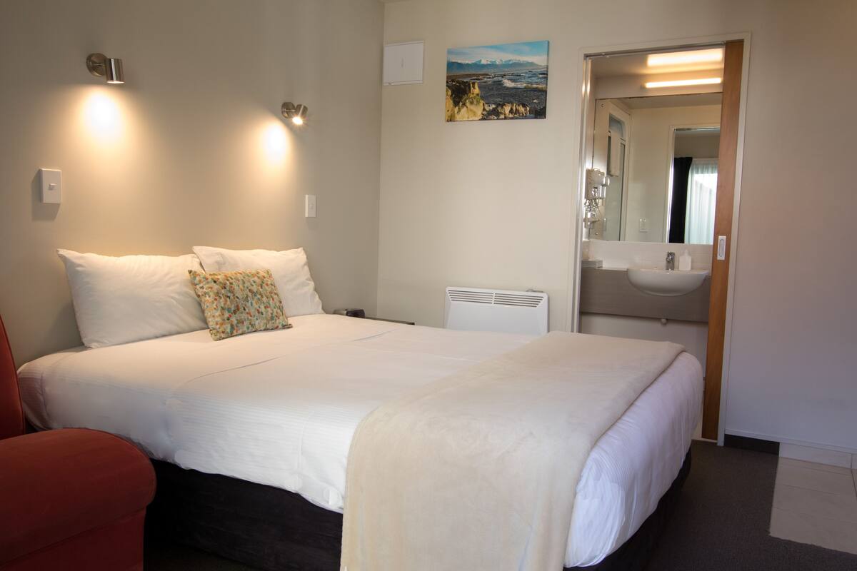 A hotel room with a bed, a red chair, a painting, and a bathroom visible through an open door.
