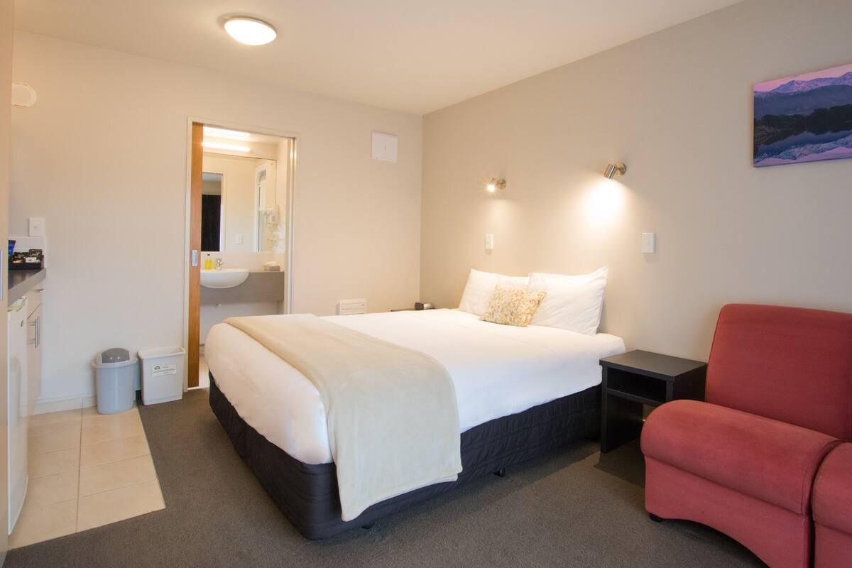 A hotel room with a bed, a nightstand, a red chair, a painting on the wall, and a bathroom visible through an open door.