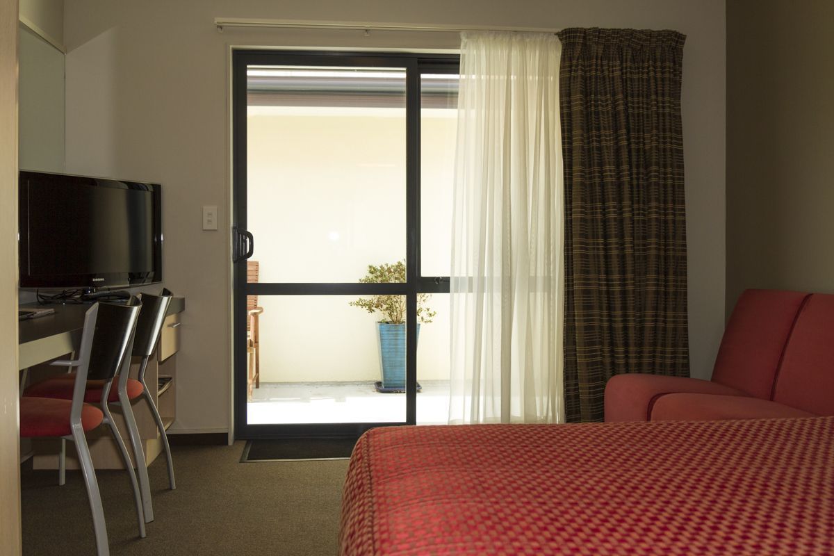 A hotel room with a bed, a desk with a TV, and a view through a sliding door to an outdoor area with a plant.