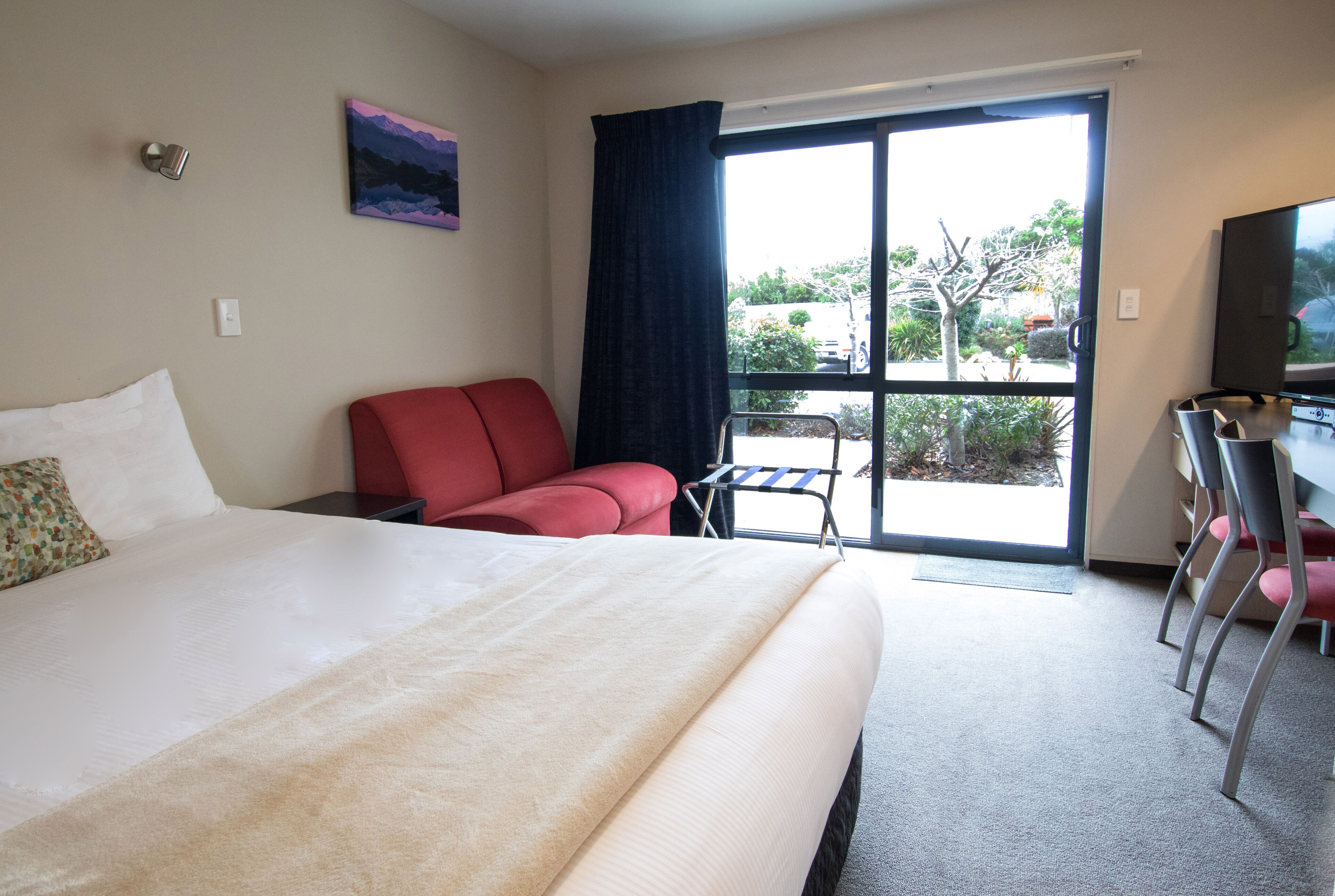 A hotel room with a large bed, a red chair, a small table, and a view of the outdoors through a sliding door.