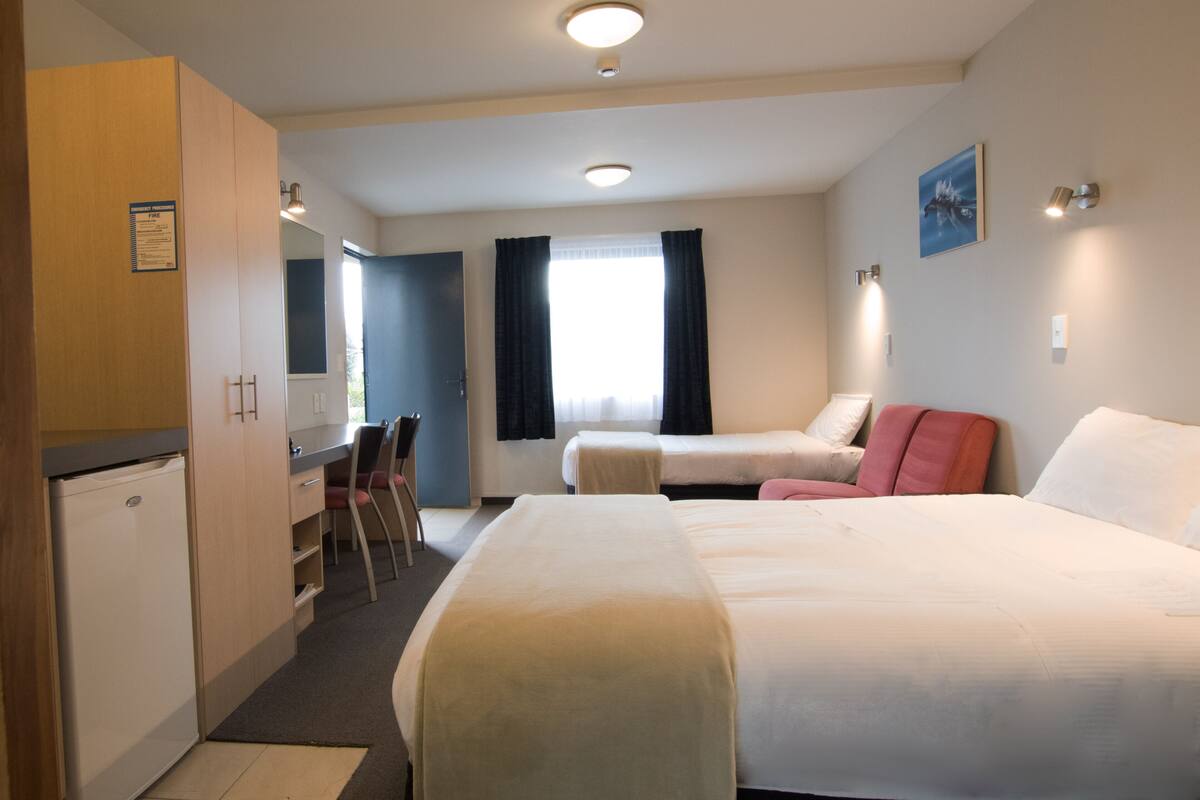 A hotel room with a bed, a desk, a chair, a small kitchenette, and a window with curtains.
