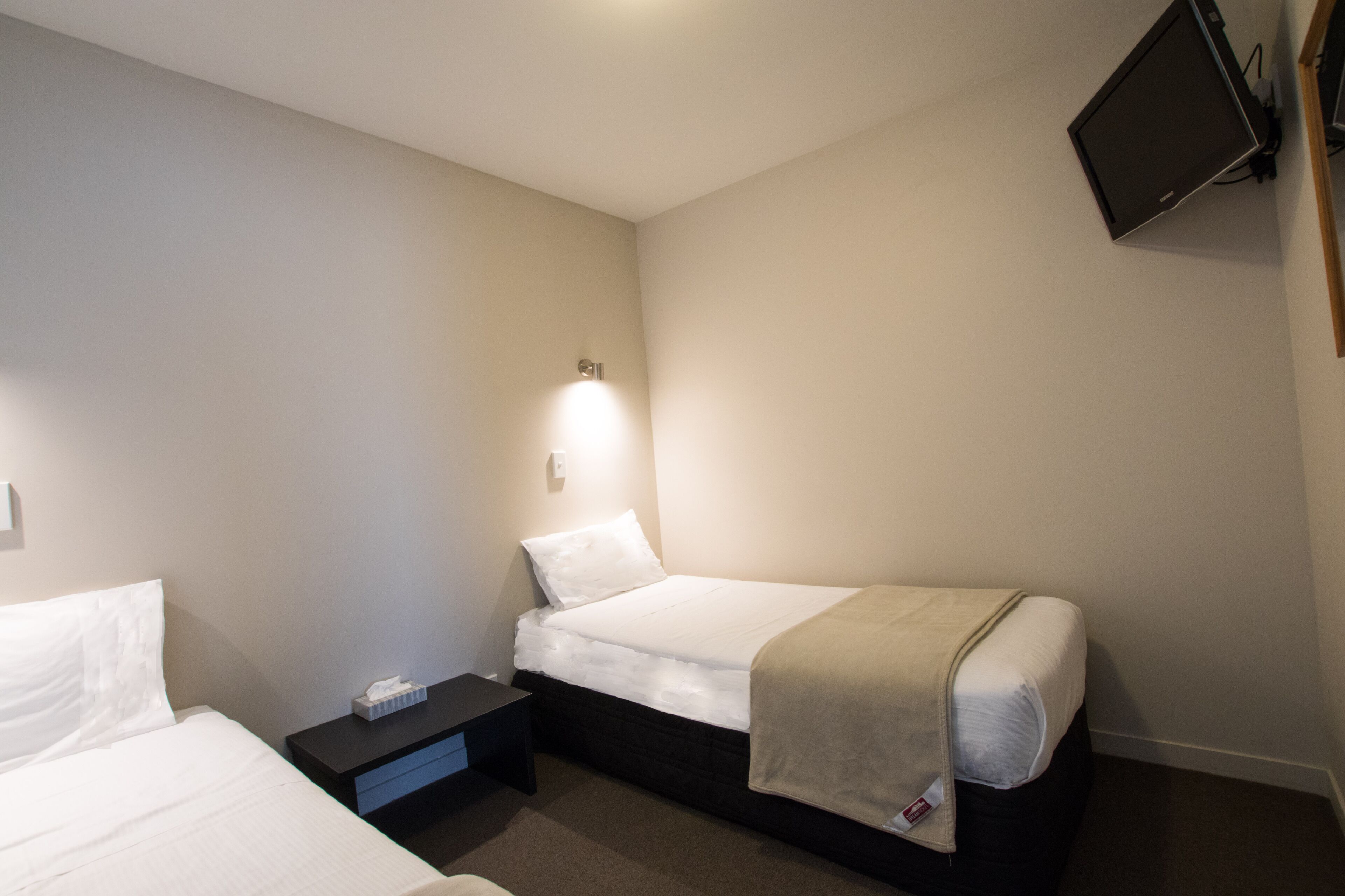 A small, neatly arranged bedroom with a single bed, a bedside table, and a wall-mounted TV.