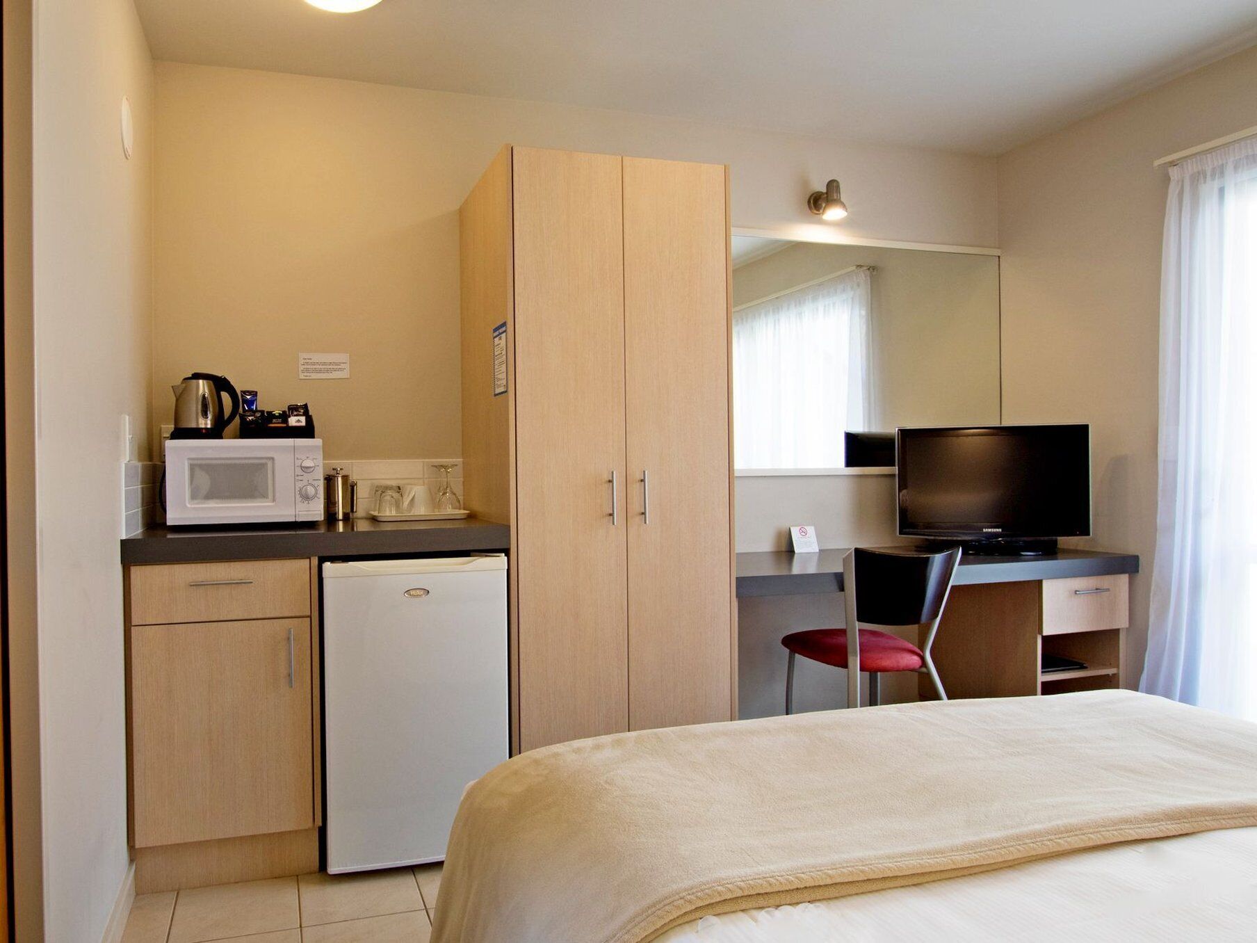 A compact hotel room with a kitchenette, a bed, a desk with a TV, and a chair.