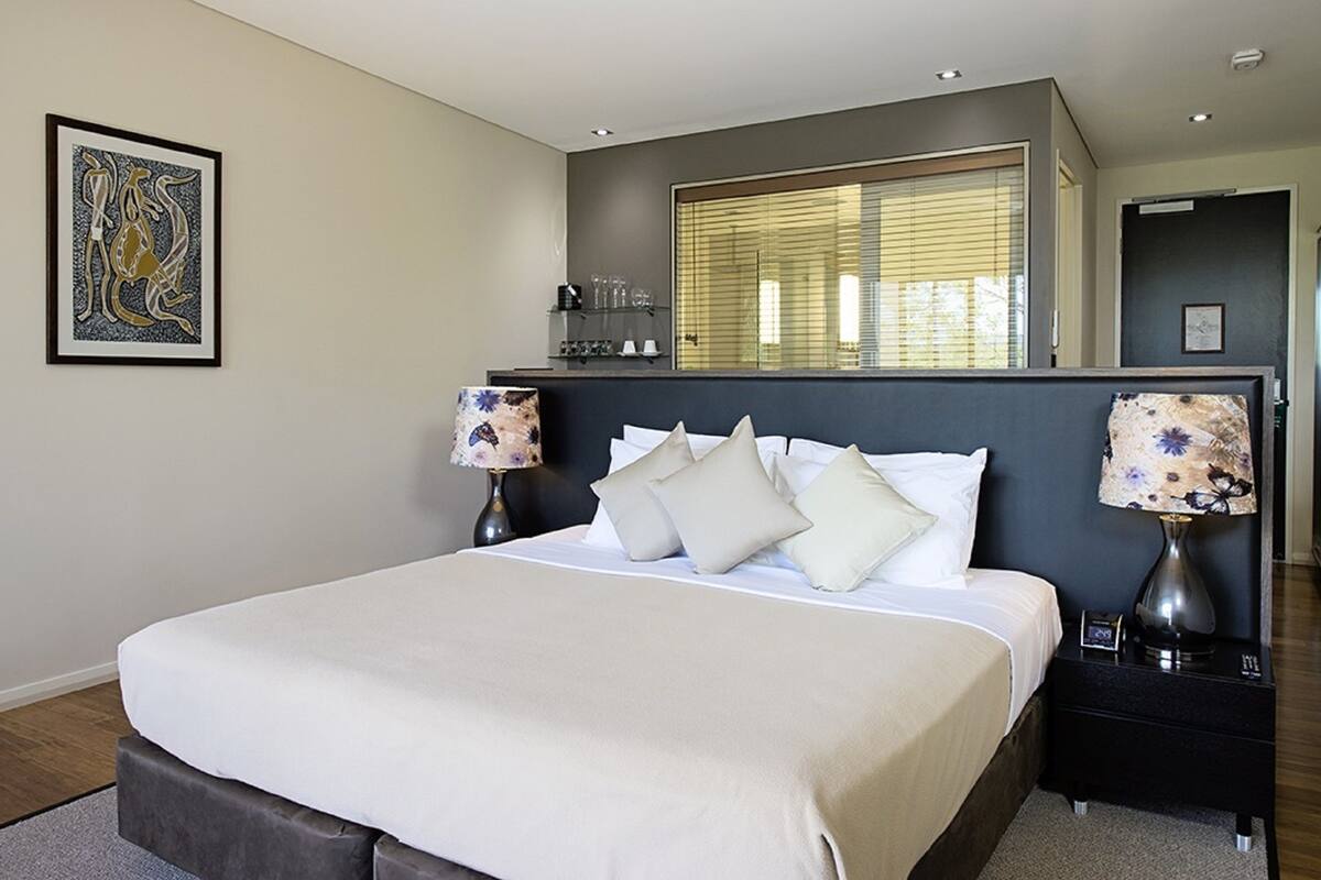 A modern bedroom with a large bed, two bedside tables with lamps, a window with blinds, and a framed artwork on the wall.