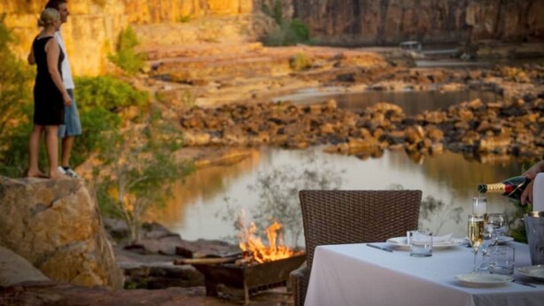 A dining area with a table set for two, overlooking a river and a fire pit. - Cicada Lodge (Nitmiluk)