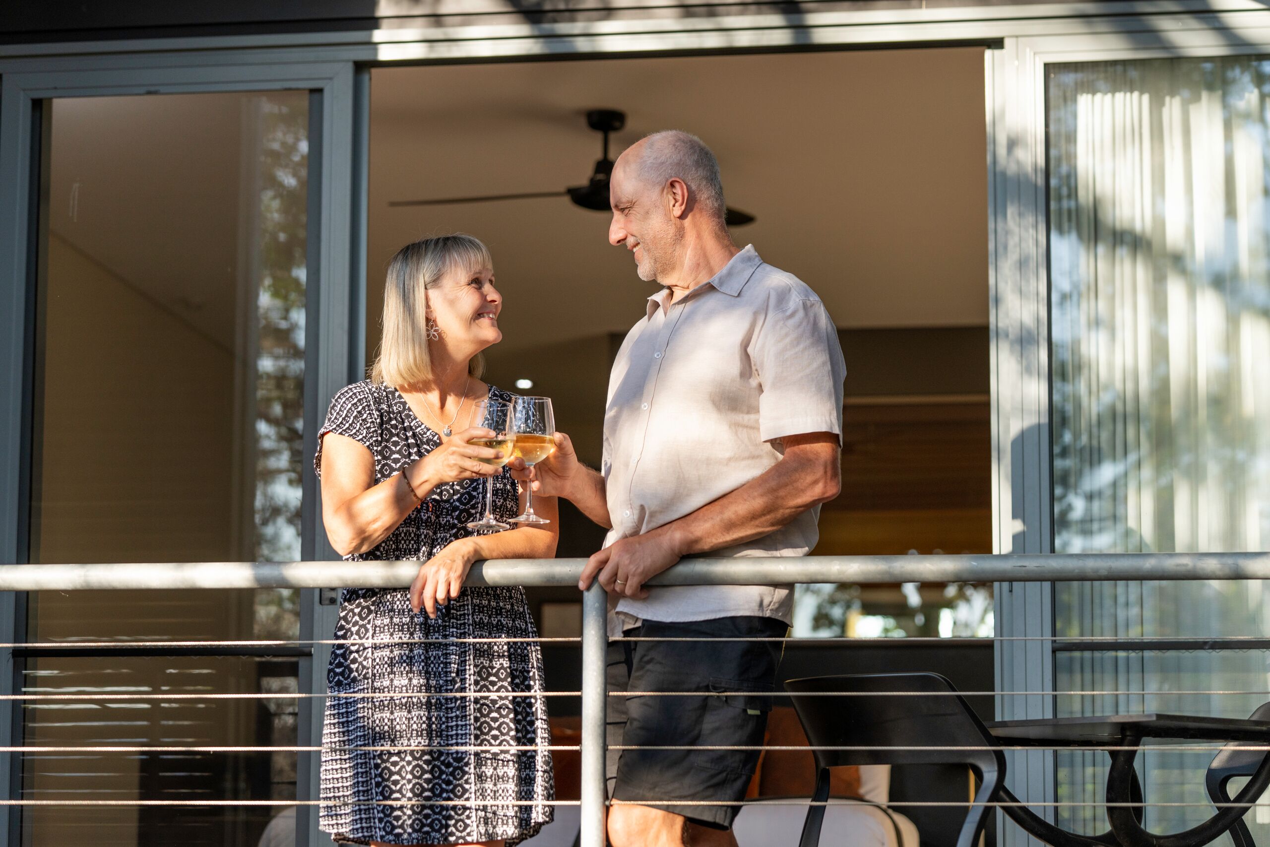 A man and a woman standing on a balcony with a railing, holding wine glasses.