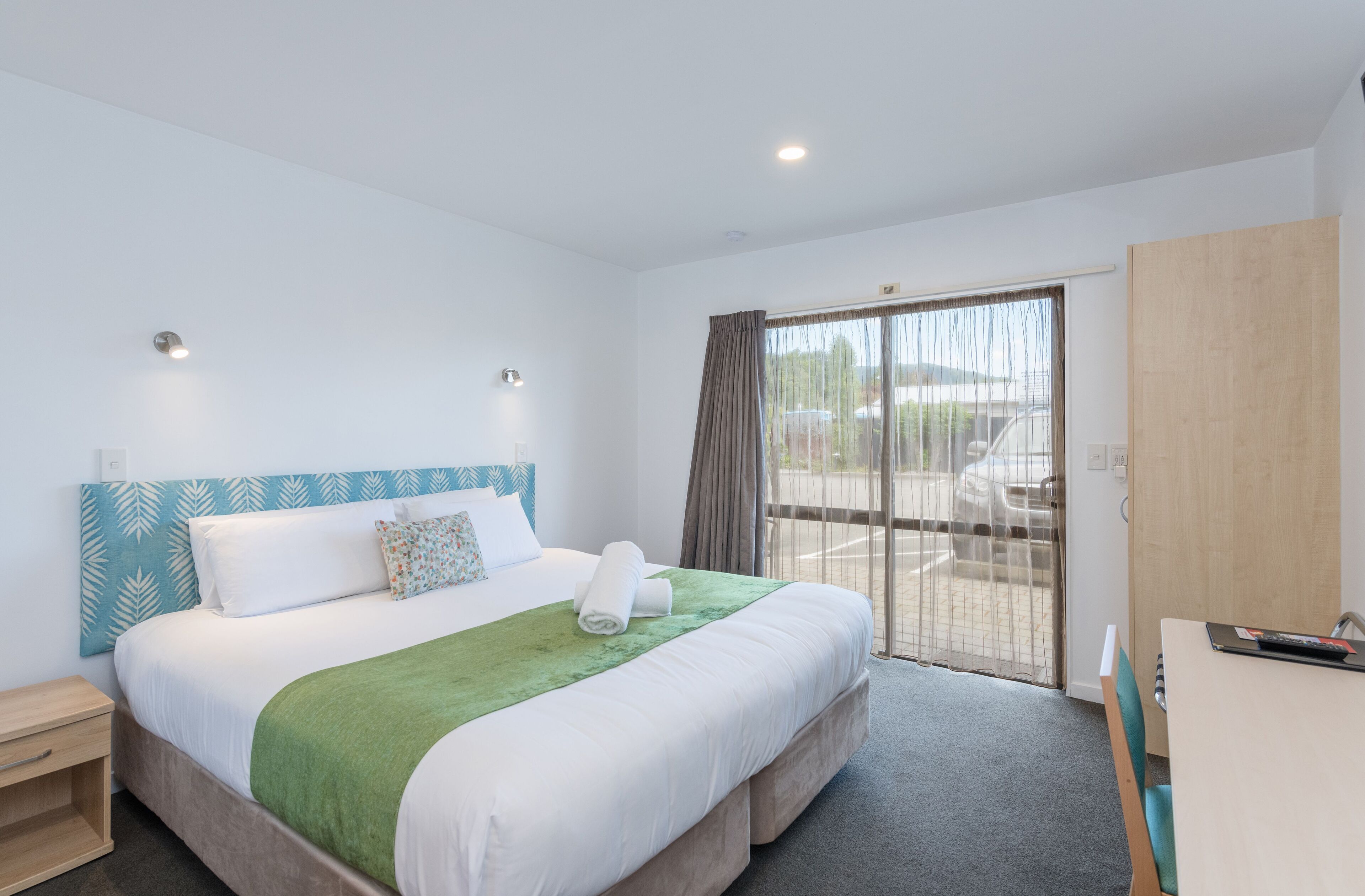 A hotel room with a large bed, a green throw blanket, a wooden headboard, a desk with a chair, and a sliding glass door to the outside.