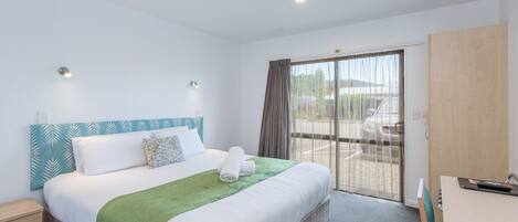 A hotel room with a large bed, a green throw blanket, a wooden headboard, a desk with a chair, and a sliding glass door to the outside.