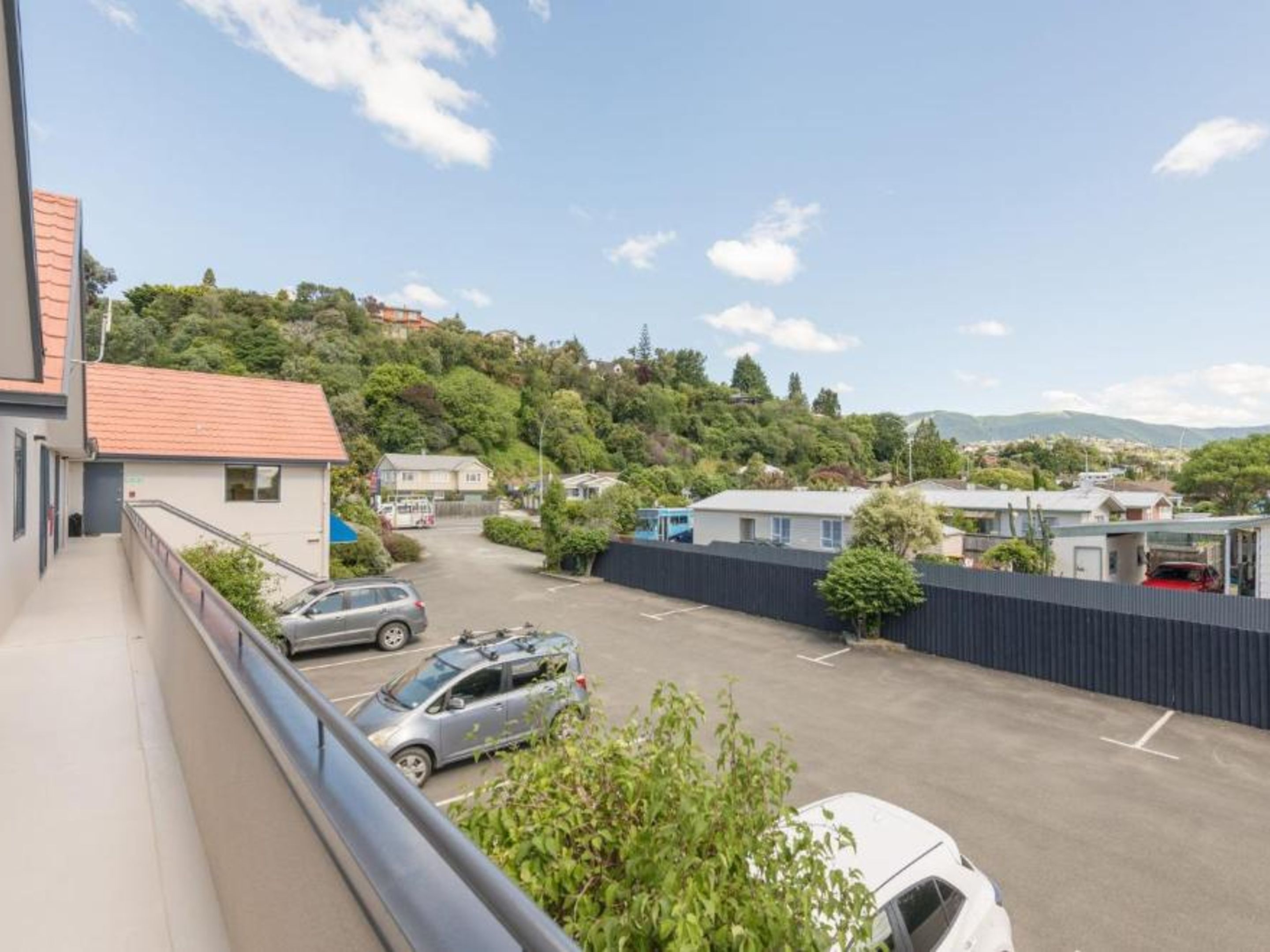 Un área de estacionamiento con varios coches, un edificio de techo rojo y una colina con árboles y casas al fondo.