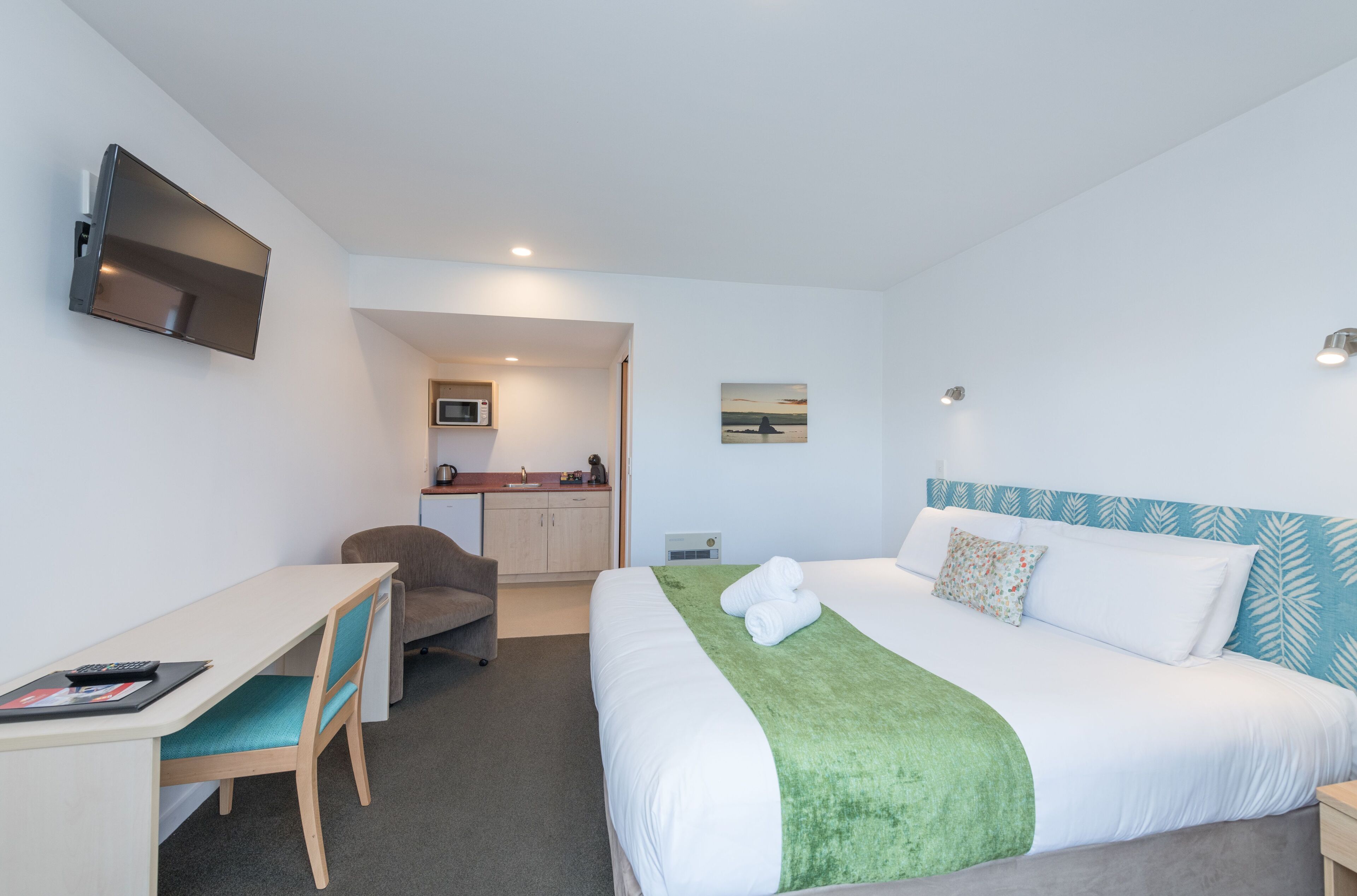 Quarto de hotel com cama, escrivaninha, poltrona, televisão, micro-ondas e um quadro na parede.