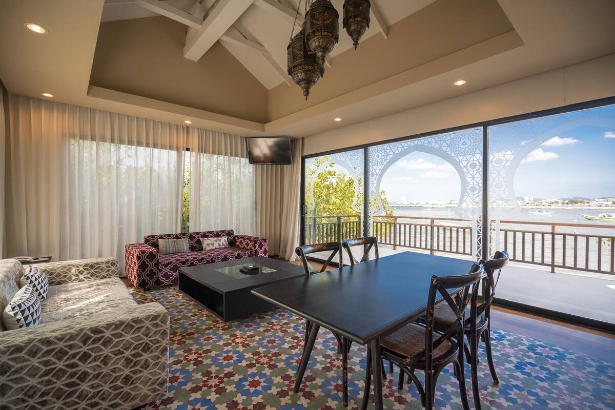 A spacious living area with a large window offering a view of the city. There is a sofa set, a coffee table, and a dining table with chairs.