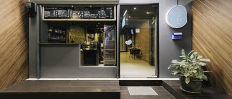 Entrada a una cafetería moderna con un cartel de menú, un botón pulsador y una planta en maceta.