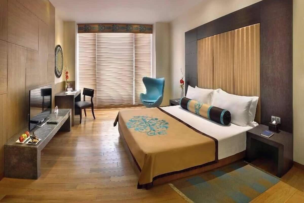 Ein Hotelzimmer mit einem großen Bett, einem Schreibtisch mit Stuhl, einem blauen Sessel und einem Fenster mit Jalousien.