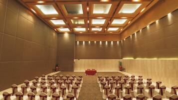 Un salón de conferencias con filas de sillas blancas, una mesa roja al frente y un techo alto con tragaluces.